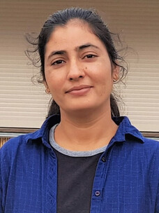 Kavita Mehlawat mit zurückgebundenem Haar und ruhigem Blick vor heller Wand.